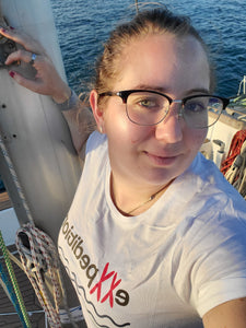 Lindsey, a white woman with blonde hair, wearing a white t-shirt that reads eXXPedition, leaning against the mast of a boat