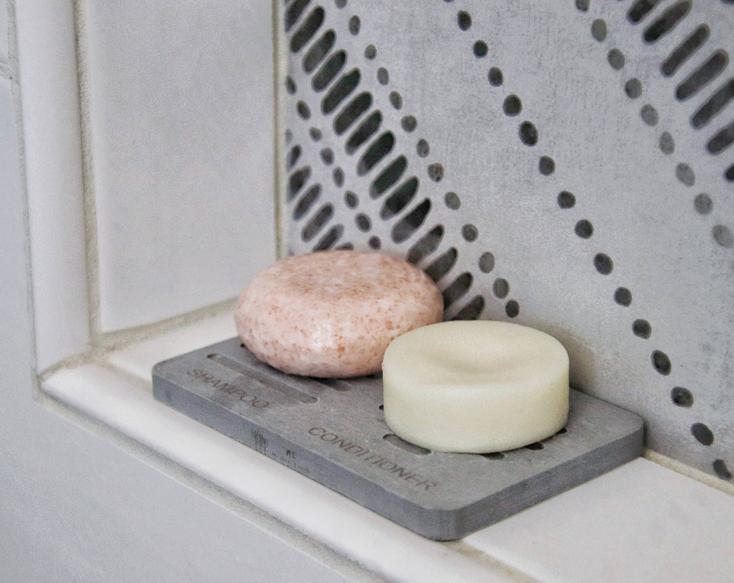 A grey dish soap holder is marked with shampoo and conditioner slots. There are bar shaped products on top.
