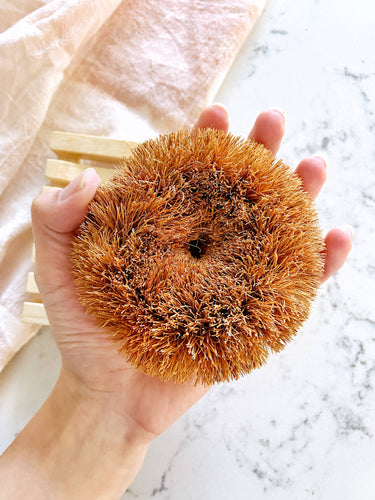 A white hand holds a round, brown coconut scrubber. 