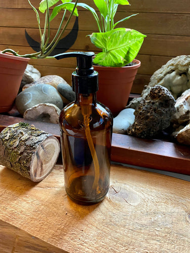 An amber glass bottle with black pump rests on a counter in front of plants and rocks.
